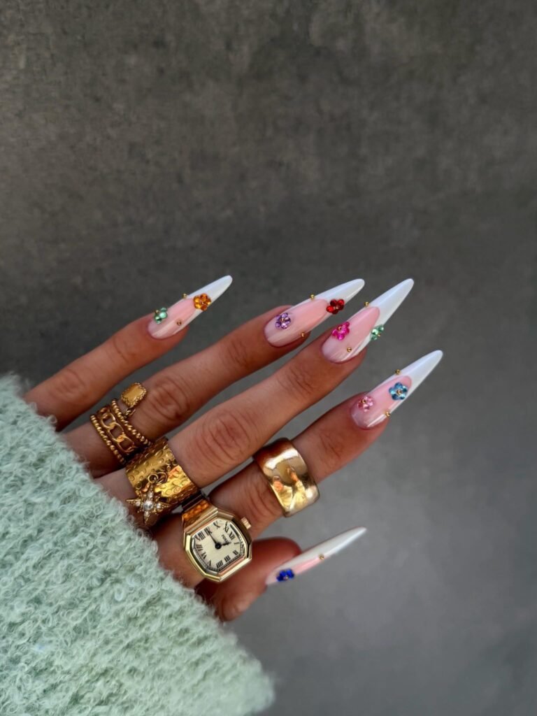 Long white stiletto nails with pastel pink bases and colorful flower gems.  