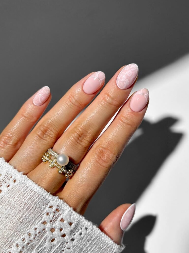 Pale pink nails with delicate lace pattern, sunlight, and pearl ring.