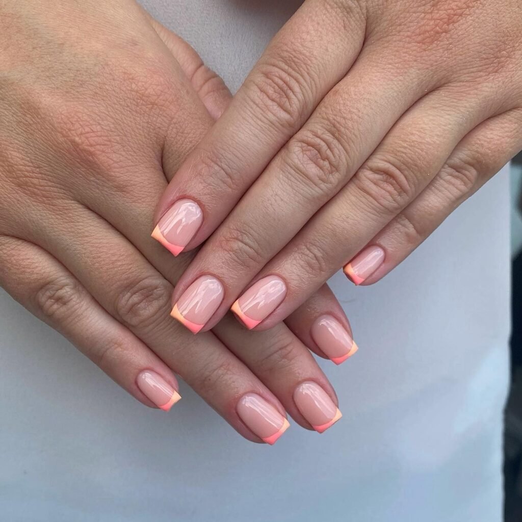 Hands with glossy nude nails and coral French tips on short squares. 