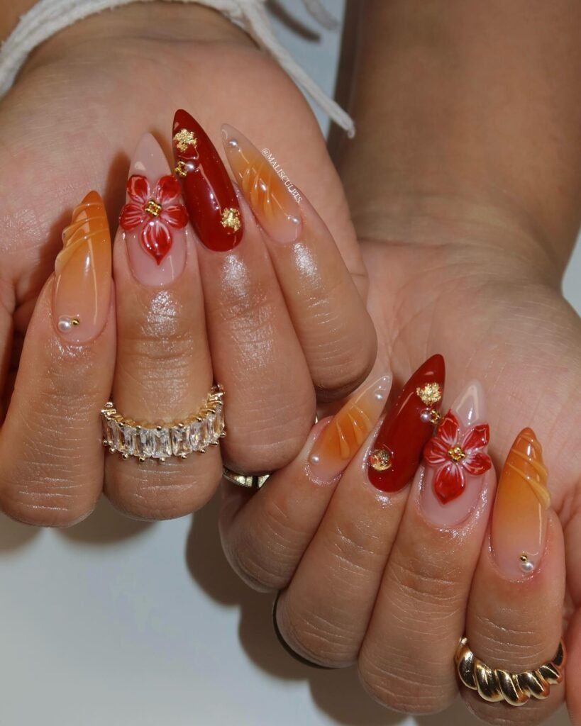 Long almond nails with red, orange, and nude tones, floral accents, gold details, and a sophisticated glossy finish.