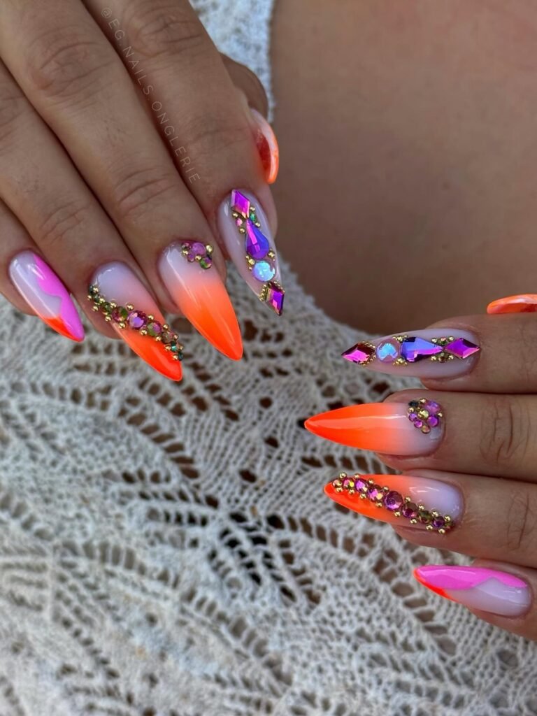 Stiletto nails with neon orange ombré tips, purple rhinestones, and gold bead accents on a lace background.
