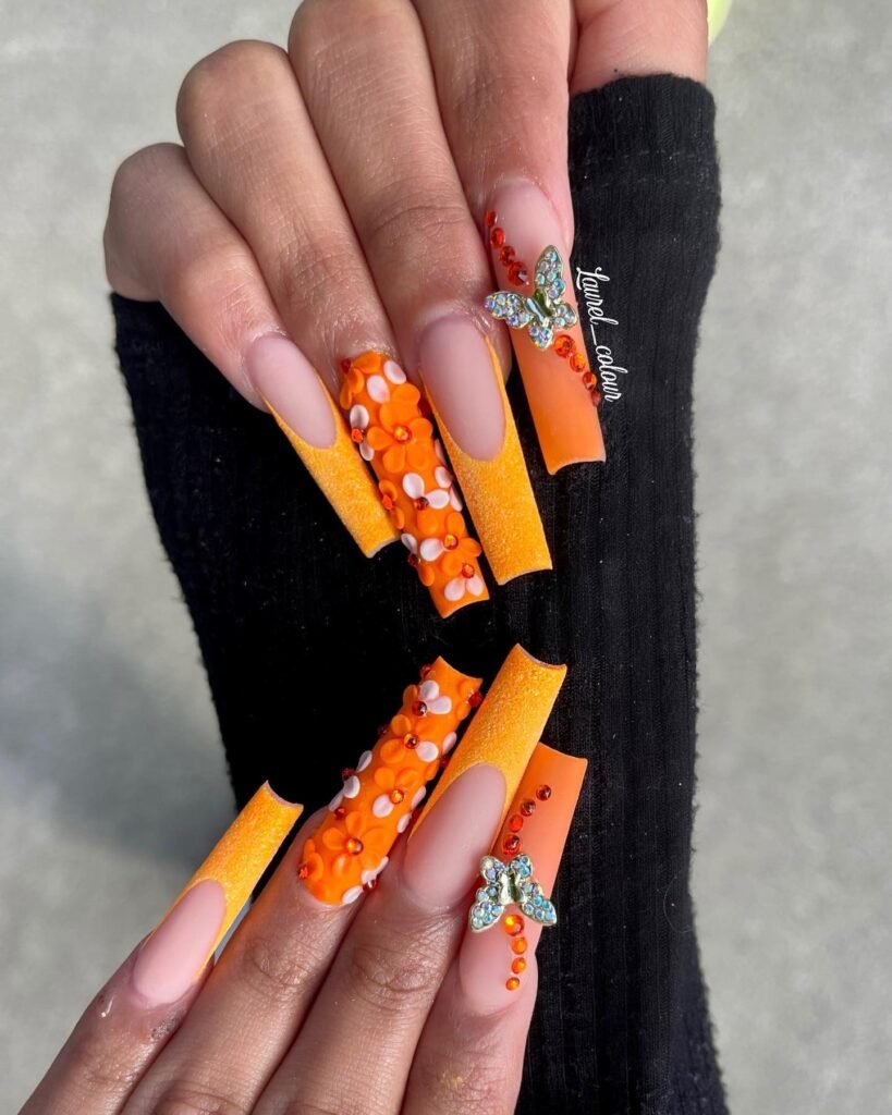 Long square orange nails with sparkly gradients, butterfly charms, rhinestone accents, and a glossy luxe finish.
